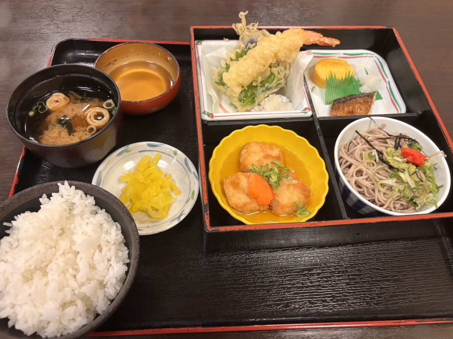 四つ仕切りのお弁当箱に海老天ぷら、そば、揚げ出し豆腐、焼き魚、卵焼きが入った極楽弁当と、ごはん・味噌汁・漬物