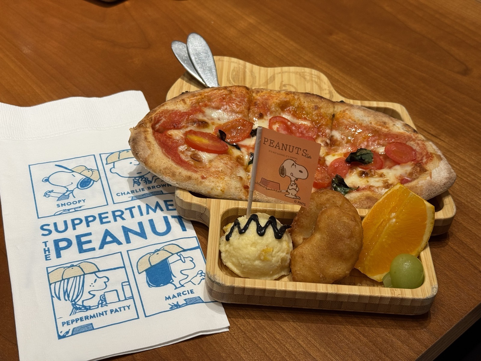 スヌーピーの顔の形をした木製プレートに、ピザ・チキンナゲット・ポテトサラダ・オレンジ・ぶどうがのっている