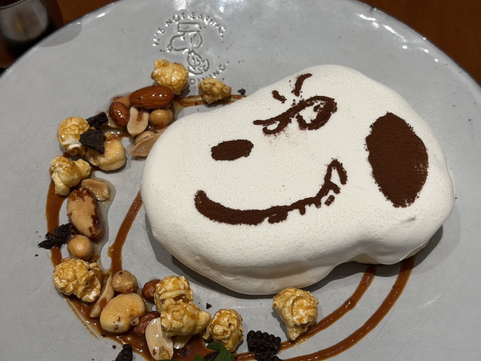 スヌーピーの顔の形のパンケーキ。キャラメルポップコーンとナッツがぐるりと添えられ、お皿にキャラメルソースが描かれている
